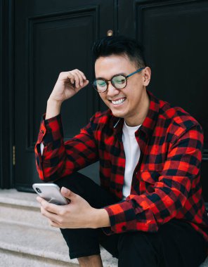 Neşeli genç Asyalı erkek öğrenci, günlük kıyafetler içinde cep telefonu kullanıyor ve bina kapısında otururken eğleniyor.