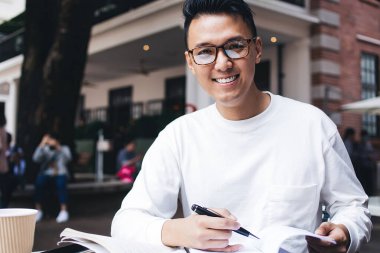 Asyalı, gözlüklü, zeki, boş not defteriyle not alan, caddedeki kafede ders çalışan ve kameraya bakan biri.