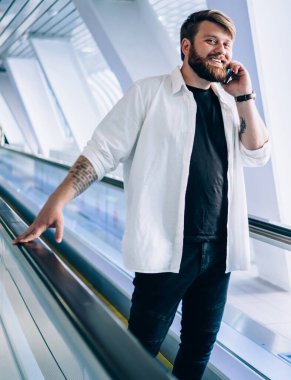 Neşeli bir erkeğin tam boy portresi havalimanı terminaline elektrikli yürüyen merdivenle gitmek ve cep telefonu cihazıyla arkadaşını aramak zamanı kontrol etmek için mutlu bir adamın cep telefonuyla iletişim kurması.