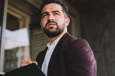 Resmi kıyafetli, düşünceli İspanyol bir adam modern binanın dışında durup planlama yaparken başka yöne bakıyor ve düşünüyor.