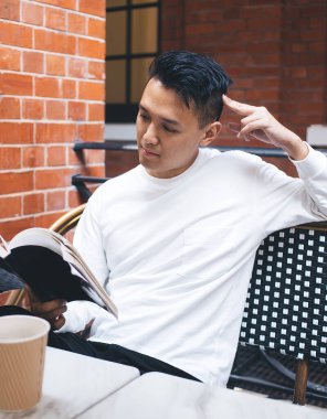 Beyaz gömlekli düşünceli, modern Asyalı bir erkek bir fincan kahveyle kafede oturuyor ve favori yazarının heyecan verici dedektif hikâyesini okuyor.