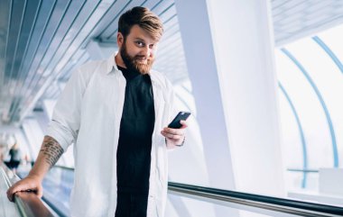 Modern cep telefonu zımbırtısıyla çevrimiçi sohbet etmek için boş zamanlarında elektrikli merdivenlerde dikilip kameraya bakan, şık giyinmiş, modern çağdan kalma hippi bir adamın yarım boy portresi.