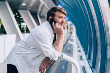 Hafif geometrik koridorda çitlere yaslanmış, cep telefonuyla konuşan hoş sakallı adamın yan görüntüsü.