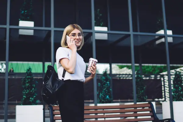 Positive young lady in casual style and backpack walking with coffee to go and calling on phone looking at camera on street design building background
