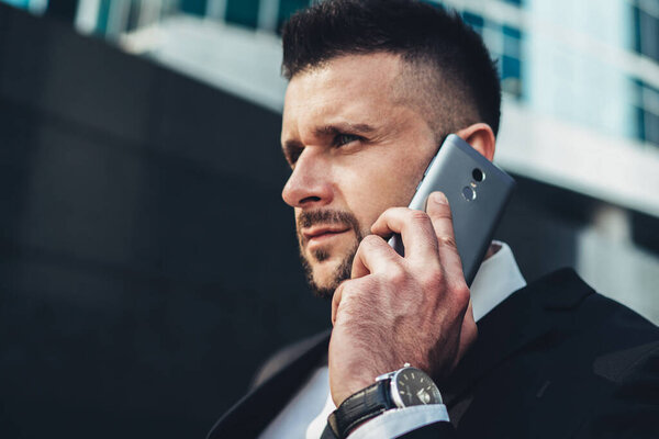 Молодой и сосредоточенный мужчина в элегантном черном костюме и смотреть разговор на смартфоне на улице в мегаполисе и смотреть в сторону