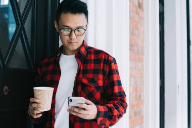 Gözlüklü, kareli tişörtlü, cep telefonu tutan ve binanın kapısında beklerken içki ısmarlayan konsantre Asyalı genç bir erkek öğrenci.