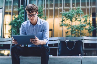 Gözlüklü yakışıklı genç bir adam. Ofis binasının önündeki bankta oturuyor. Manhattan, New York 'ta bir sokakta tablet kullanıyor.