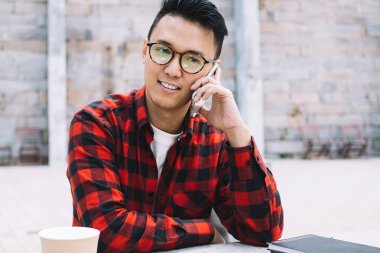 Düşünceli, genç, düşünceli, gözlüklü ve kareli tişörtlü Asyalı adam dışarıda, şehir masasında telefonla konuşurken başka tarafa bakıyor.