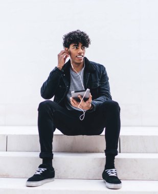 Tabletle merdivenlerde oturmuş kulaklıkla müzik dinlerken beyaz duvara bakan Afro-Amerikalı genç yakışıklı bir adam.
