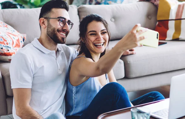 Happy sweet couple in casual wear relaxing and cuddling by modern couch while shooting selfie and using laptop in apartment with contemporary interior