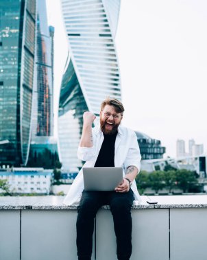 Beyaz gömlekli neşeli genç sakallı adam başarılı bir açılışı kutluyor iş bölgesinde çitlerde oturuyor.