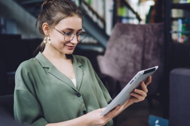 Günlük kıyafetleri ve modaya uygun gözlükleri olan neşeli genç bayan evde oturup tablet izliyor ve video seyrediyor ve gülümsüyor.