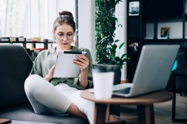 Serbest giyinen genç ve güzel bir bayan kütüphanede koltukta oturup tablet kullanarak internetten bilgi arıyor.