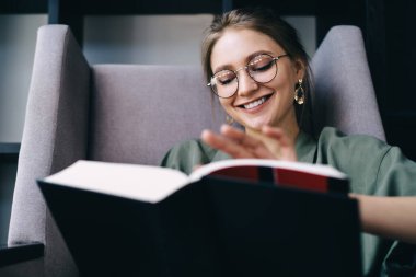 Gözlüklü neşeli bayan öğrenci kitaplıkların yanındaki modern kütüphanenin sandalyesinde otururken siyah kapaklı büyük bir kitap okuyor.