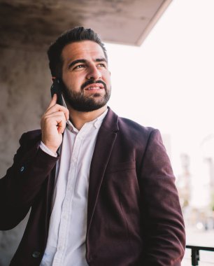Düşünceli İspanyol takım elbiseli adam, modern şehirde çalışırken terasta akıllı telefon konuşmaları yaparken başka tarafa bakıyor ve düşünüyor.
