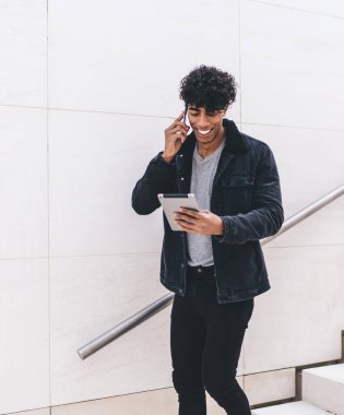 Modern stil güler yüzlü genç esmer Afrikalı Amerikalı adam telefonda konuşuyor ve tablet kullanıyor. Gün ışığında şehir merdivenlerinde dikilirken.