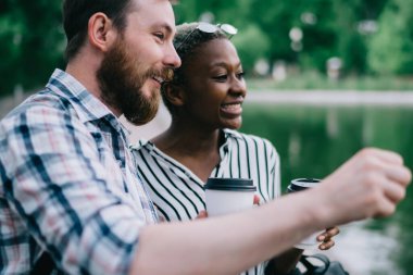 Genç, çoklu etnik çeşitliliğe sahip kadın ve erkek kahve içerek ve gün ortasında gölün kenarında komik sohbetler yaparak eğleniyorlar.