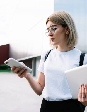 Gözlüklü, günlük giysiler içinde dikkatli bir kadın. Ofis binasının yanındaki caddede dikilirken konsantrasyonuyla akıllı telefona göz atıyor.