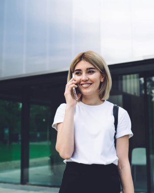 Neşeli nedensel modern sarışın kadın siyah sırt çantalı sokak binasına karşı yürürken telefonla konuşuyor ve mutlu bir şekilde gülümsüyor.
