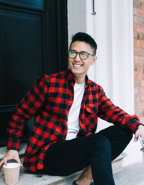 Tatmin olmuş Asyalı ekose gömlekli ve gözlüklü genç bir adam. Kapıda vakit geçirip kahvenin tadını çıkarıyor ve gülümsüyor ve başka tarafa bakıyor.