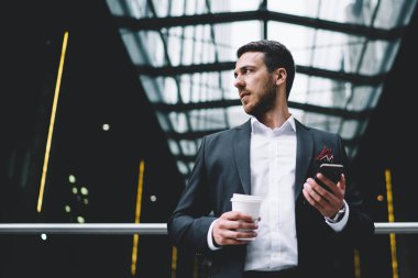 Resmi takım elbiseli, sakallı bir erkek müdürün elinde cep telefonuyla modern ofis binasına bakıyor.