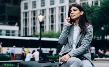 Gri formalı oldukça çekici bir kadın taştan bir bankta oturup telefonda konuşurken yüzünü çeviriyor.