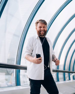 Kolları kulaklıklı, beyaz tişörtlü mutlu sakallı adam yaya köprüsünde akıllı telefondan müzik dinliyor.