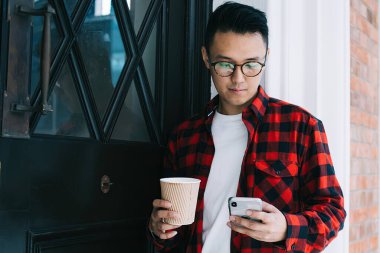 Gözlüklü, kareli tişörtlü, cep telefonu tutan ve binanın kapısında beklerken içki ısmarlayan konsantre Asyalı genç bir erkek öğrenci.