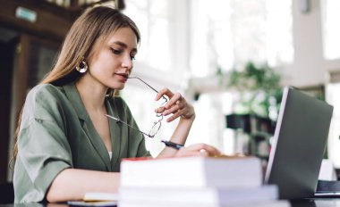 Uzun saçlı, uzun gözlüklerini ısıran düşünceli genç bir kadının yan görüntüsü. Dizüstü bilgisayar üzerinde çalışırken açık kütüphanede oturuyor.