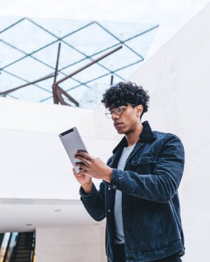 Kot takım elbiseli, tabletli gözlüklü, geniş bir odada duran ve sosyal ağlarda sohbet eden dalgın kıvırcık siyah adamın yan görüntüsü.