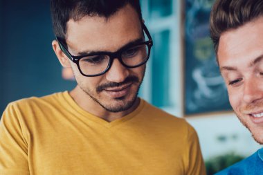 Keyifli genç adam, günlük giysiler içinde, kafeteryadaki akıllı telefon ekranını gösteren gözlüklü yakışıklı arkadaşıyla rahatlıyor.