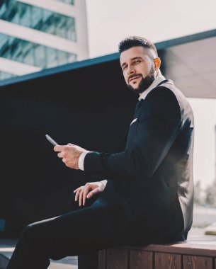 Siyah takım elbiseli, cep telefonuyla gezen şık bir erkeğin yan görüntüsü. Şehirde, güneş ışığında ahşap bir podyumda oturuyor.