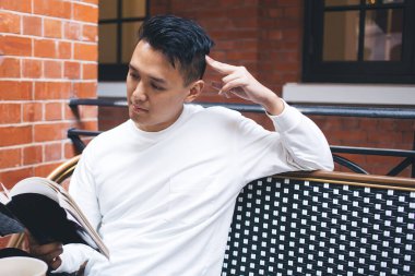 Beyaz gömlekli düşünceli, modern Asyalı bir erkek bir fincan kahveyle kafede oturuyor ve favori yazarının heyecan verici dedektif hikâyesini okuyor.