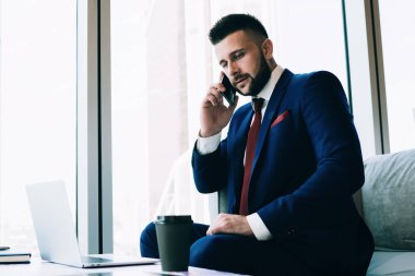 Takım elbiseli sakallı bir erkek, telefona cevap veriyor ve güncel ofise gitmek için laptop ve içeceklerle masanın yanındaki koltukta otururken düşünüyor.