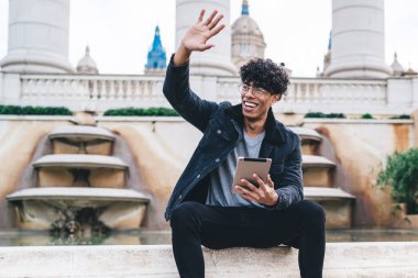 Siyah elbiseli genç Afro-Amerikan erkek öğrenci çeşmenin kenarında oturmuş, elinde tabletle kentin arka planında el sallıyor.