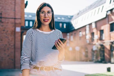 Teknoloji donanımlı neşeli kız öğrencilerin yarım boy portresi. Açık havada durup kameraya gülümsüyor. Cep telefonuyla 4G 'ye bağlanan mutlu hippi kız.