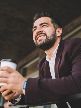 İspanyol patronun içki içme açısı düşük ve modern akıllı telefon diğer tarafa bakıyor ve modern bina terasına yaslanırken gülümsüyor.