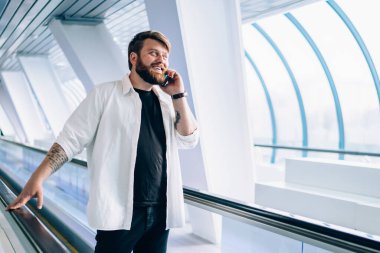 Siyah tişörtlü ve beyaz tişörtlü hoş sakallı adam cep telefonuyla konuşuyor hafif geometrik tünelde tırabzan tutuyor.