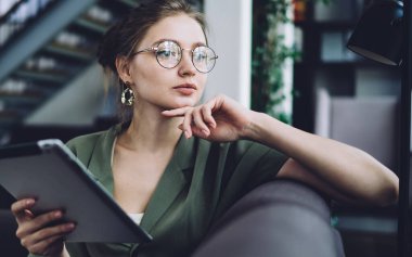 Yuvarlak bardaklı düşünceli genç bayan koltukta oturup çene okşuyor ve ofisteki kanepede tabletle otururken düşüncelerini başka yöne çeviriyor.