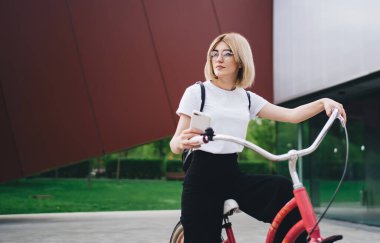 Gözlüklü, kendine güveni olan, kırmızı bisikletli ve sosyal medyada gezinen akıllı telefonlu, gözünü çevirmiş bir kadın.