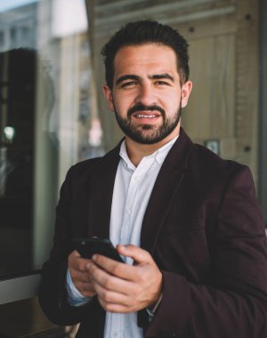 Sakallı, takım elbiseli İspanyol patron pencereye yaslanırken kameraya bakıyor ve modern binanın dışındaki akıllı telefona bakıyor.