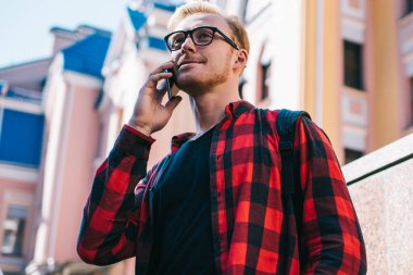 Kırmızı sakallı, gözlüklü, kırmızı ve siyah tişörtlü bir erkek yazın şehirde cep telefonundan arama yapıyor.