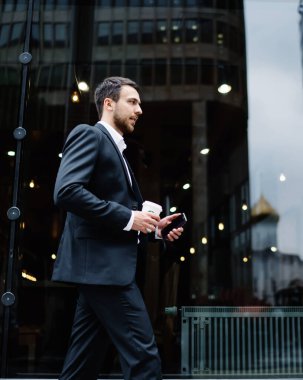 Şehir merkezindeki şehir ışıklarını yansıtan siyah camlar boyunca yürüyen koyu renk, şık takım elbiseli ve kahveli kendine güvenen işadamının yan görüntüsü.