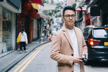 Kendine güvenen, bej ceketli ve moda gözlüklü Asyalı erkek gazeteci şehir caddesinde yürürken fotoğraf makinesiyle haber yapıyor ve kameraya bakıyor.