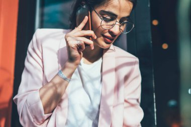 Gözlüklü, resmi kıyafetleri olan, ahşap bankta otururken telefonda konuşan ve şehir merkezindeki bulanık renkli çağdaş binaya karşı derin düşüncelere dalmış bir iş kadınından.
