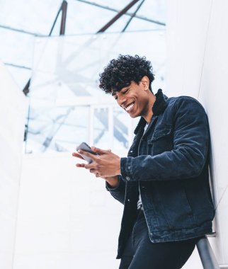 Beyaz modern binada dikilirken, kıvırcık saçlı, akıllı telefonuyla mesajlaşan şık giyinmiş neşeli, Afro-Amerikan bir adam.