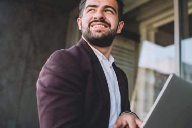 Sakallı, takım elbiseli, dizüstü bilgisayarı karıştıran ve modern binanın önünde gülümseyerek duran İspanyol bir adam.