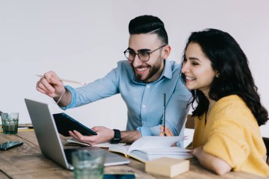 Gündelik giysiler giyen mutlu çift dizüstü bilgisayara ve tablete bakarken not alıyor ve evdeki masada oturuyor.