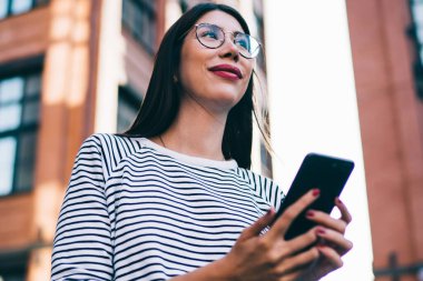 Milenyum blogcusu elinde cep telefonuyla elinde başka bir yere bakıyor ve gülümsüyor çevrimiçi iletişim ve telefon için eğleniyor, 4G kablosuz internete bağlanan pozitif hippi kız.