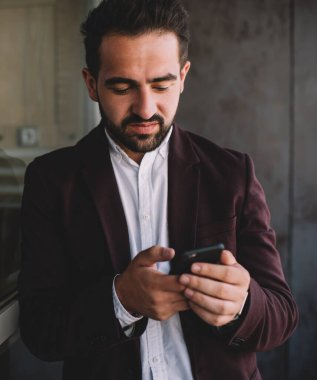 Ciddi etnik sakallı, şık takım elbiseli, pencereye yaslanmış, akıllı telefonları karıştıran ve şehrin dışındaki modern binaların önünde duran bir adam.
